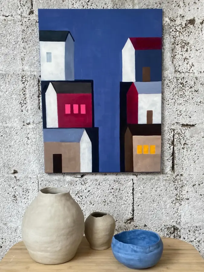 Cuadro de casas blancas y rojas sobre fondo azul colgado en una pared de bloque, con vasijas y un cuenco azul delante